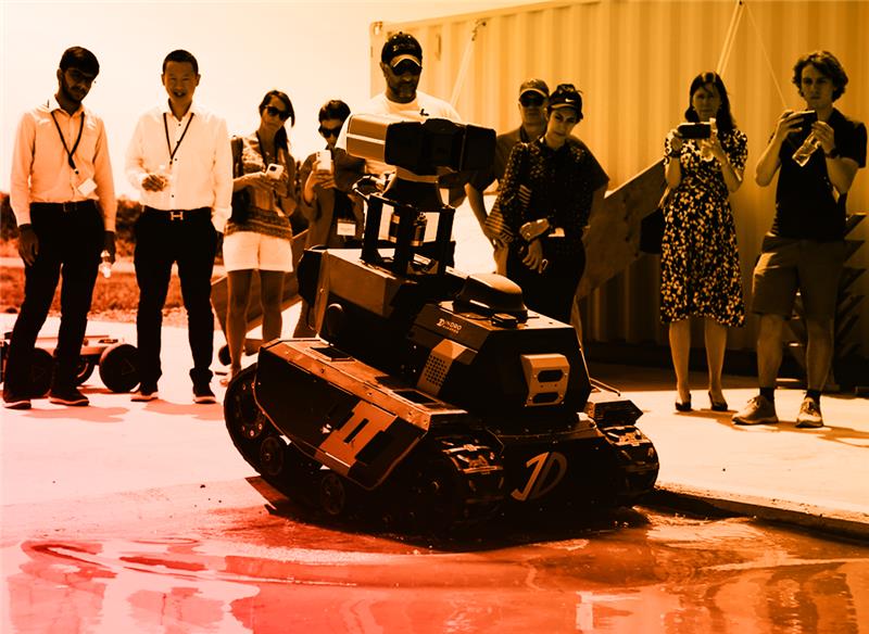 An image of a group of people watching the performance of a drone. The image has an orange gradient.